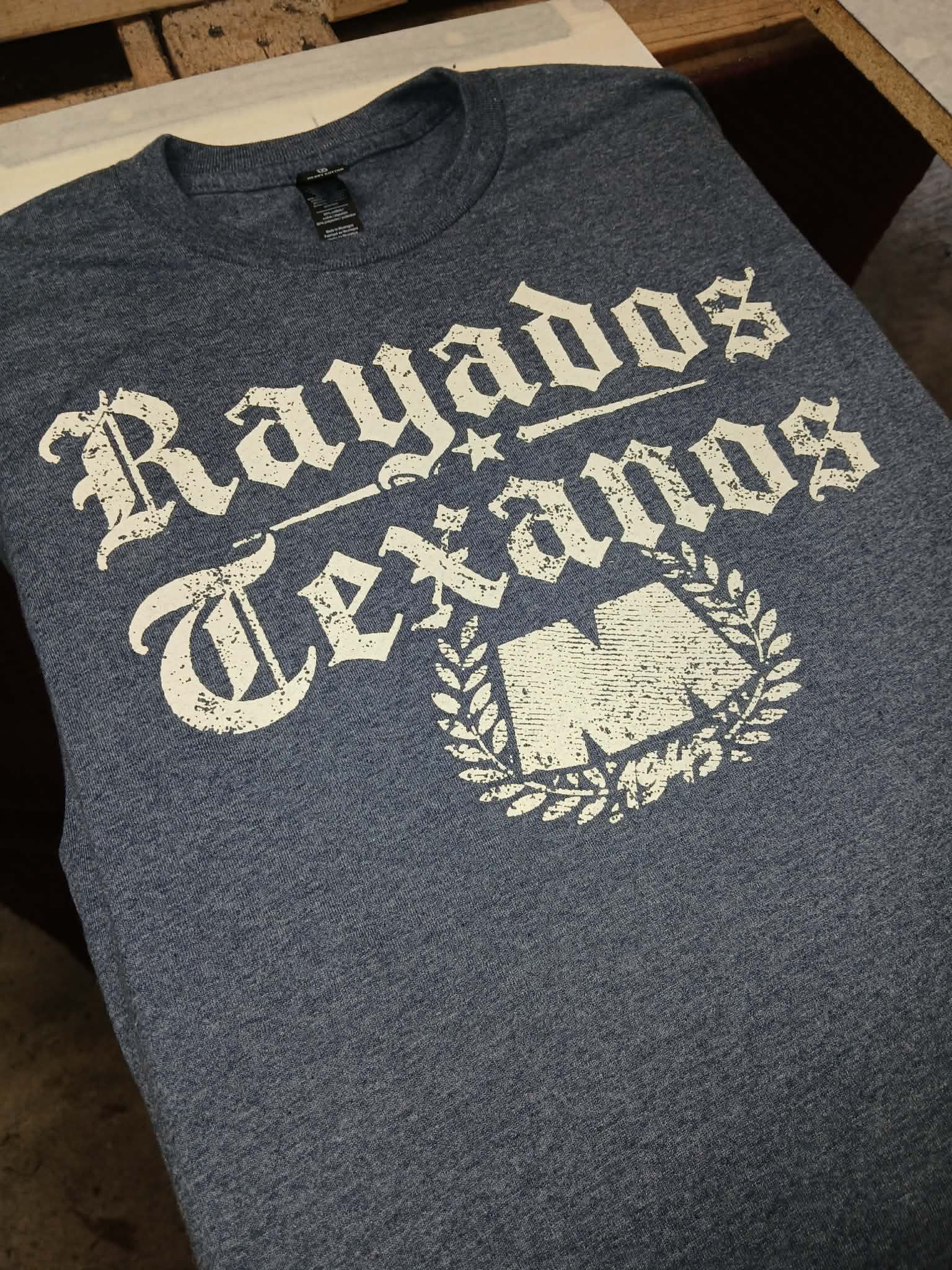 A heather navy blue t-shirt with a distressed white graphic that reads 'Rayados Texans' and features a large 'T' inside a laurel wreath, laid flat on a wooden surface.