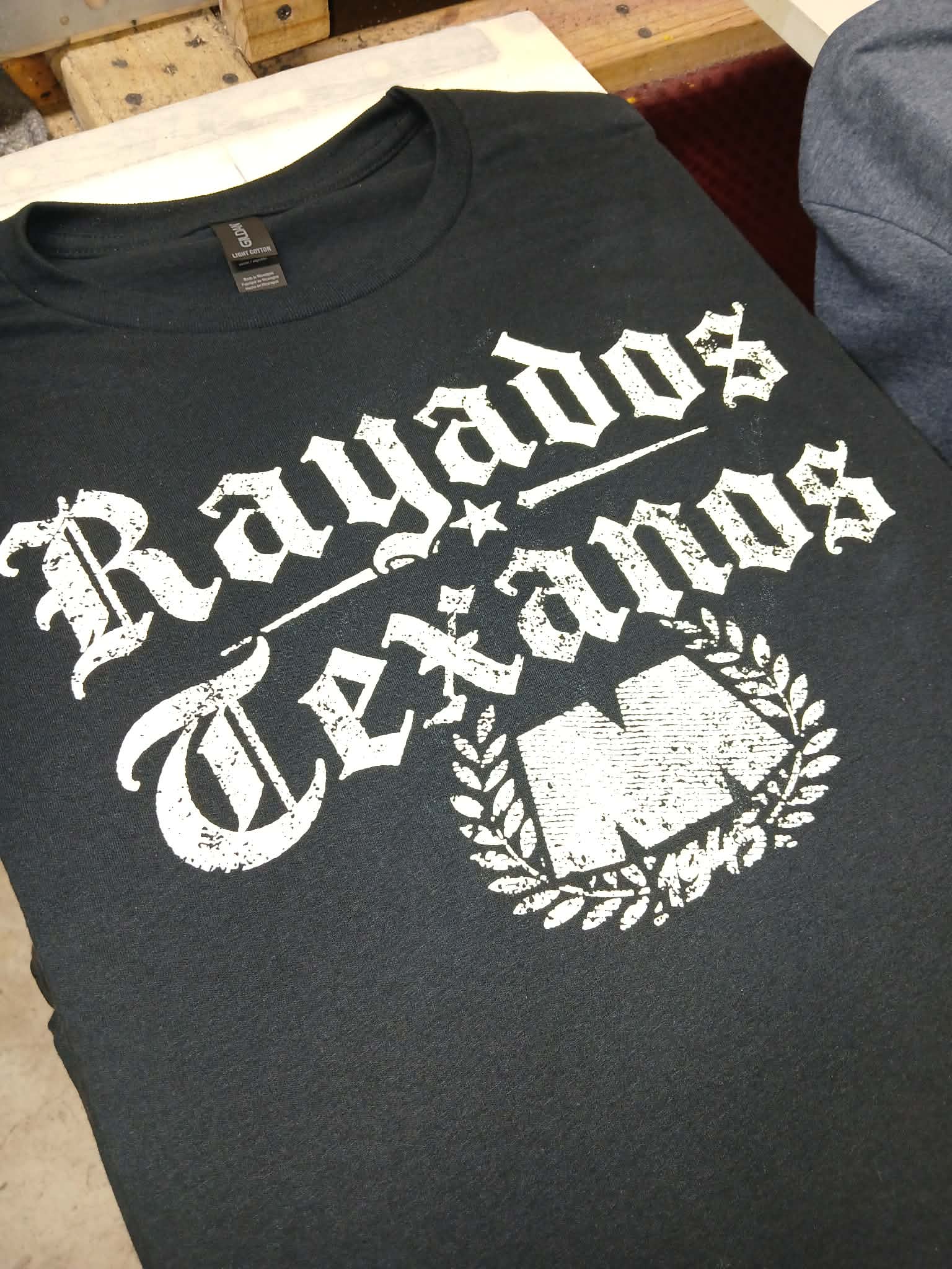 A black short-sleeve t-shirt with a distressed white graphic print reading 'Rayados Texanos' and a large 'M' inside a laurel wreath, laid flat on a light surface.