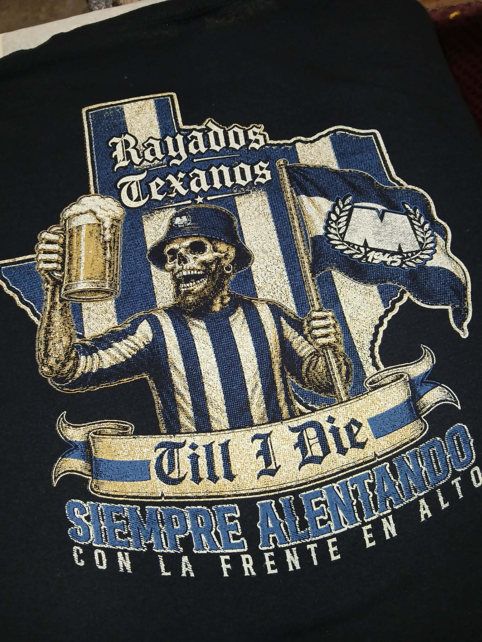 A close-up of a black t-shirt featuring a large graphic print with a skeleton referee, a beer mug, a Texas-shaped outline, and the text 'Rayados Texanos' and 'Till I Die Siempre Alientando Con La Frente En Alto'.